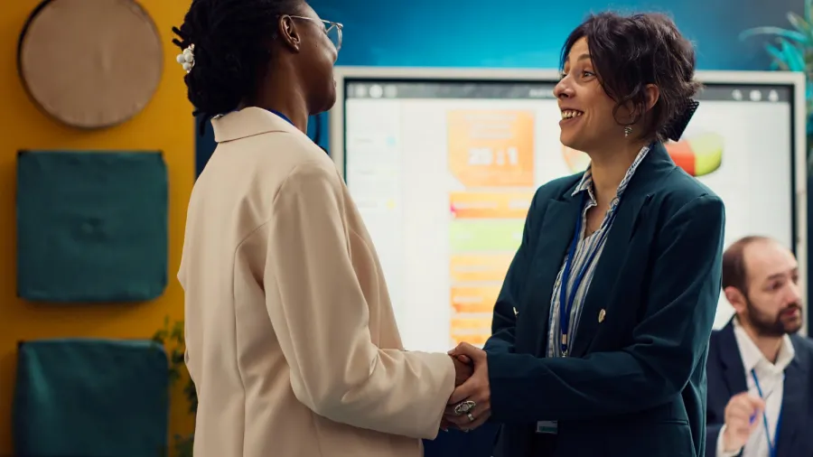 Two women wearing business attire shake hands and smile at each other in an office setting, with a man working at a desk and a presentation screen with charts in the background.
