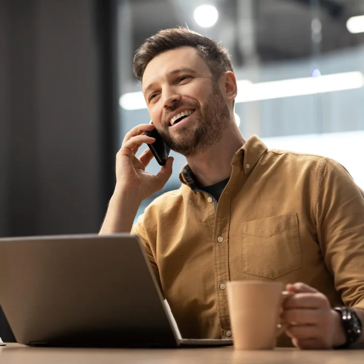 A smiling man sits at a desk with a laptop, talking on a smartphone and holding a coffee mug, in a modern office setting.