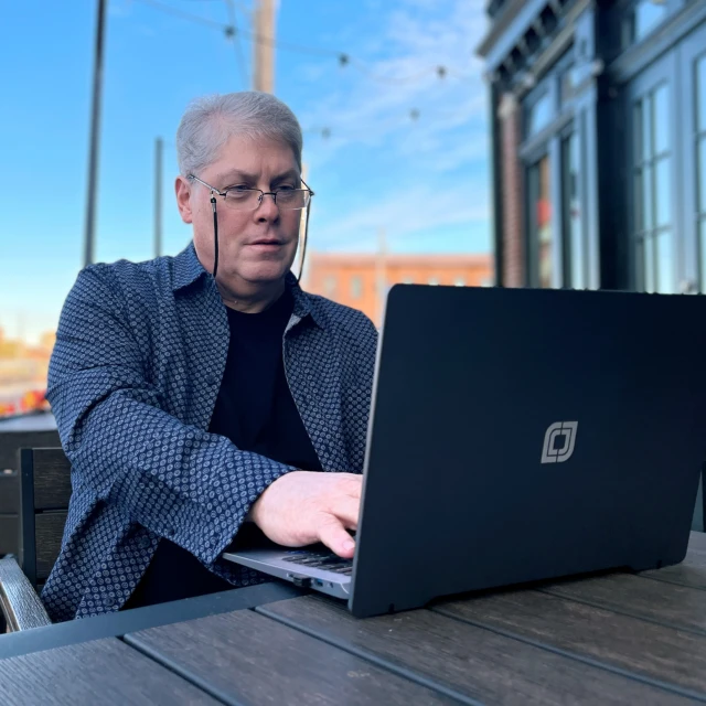 A man with gray hair and glasses sits outdoors at a wooden table, working on a laptop. He wears a patterned shirt and looks focused. Buildings and string lights are visible in the background.
